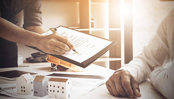 person signing a document while another person watches with small model houses on the table related to real estate deals including five essential steps